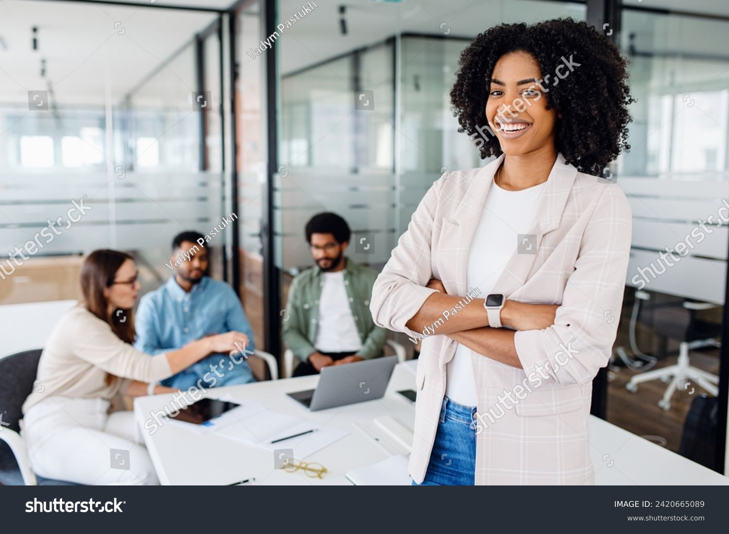 Smart high-skilled young Brazilian woman with a cheerful smile stands with arms crossed on the forefront in a modern office setting with her colleagues collaborating in the background, team synergy #2420665089