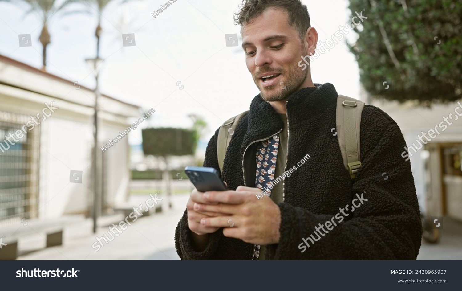 Smiling bearded young man using smartphone on a sunny city street, depicting urban life and technology. #2420965907
