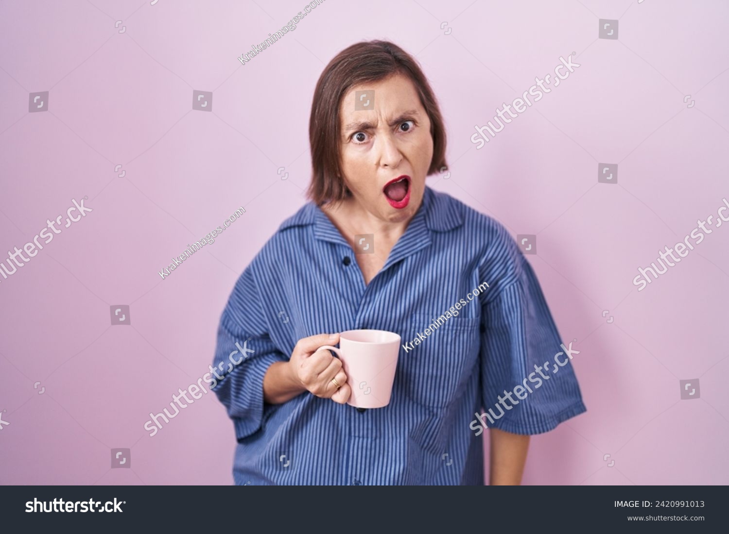 Middle age hispanic woman drinking a cup coffee in shock face, looking skeptical and sarcastic, surprised with open mouth  #2420991013