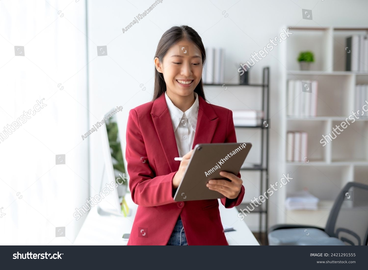 Professional young Asian businesswoman smiling while working on a digital tablet in a modern office setting. #2421291555