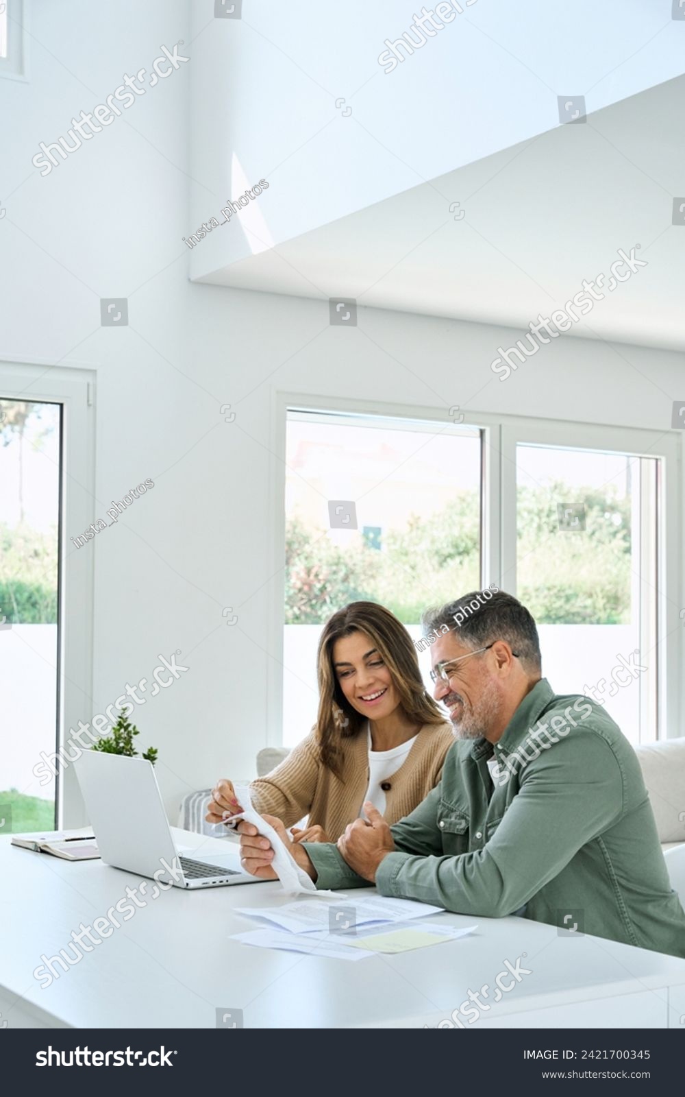 Happy mature couple calculating money expenses savings using laptop at home. Middle aged man and woman checking receipts for tax refund, paying bills online sitting at table in living room. Vertical. #2421700345
