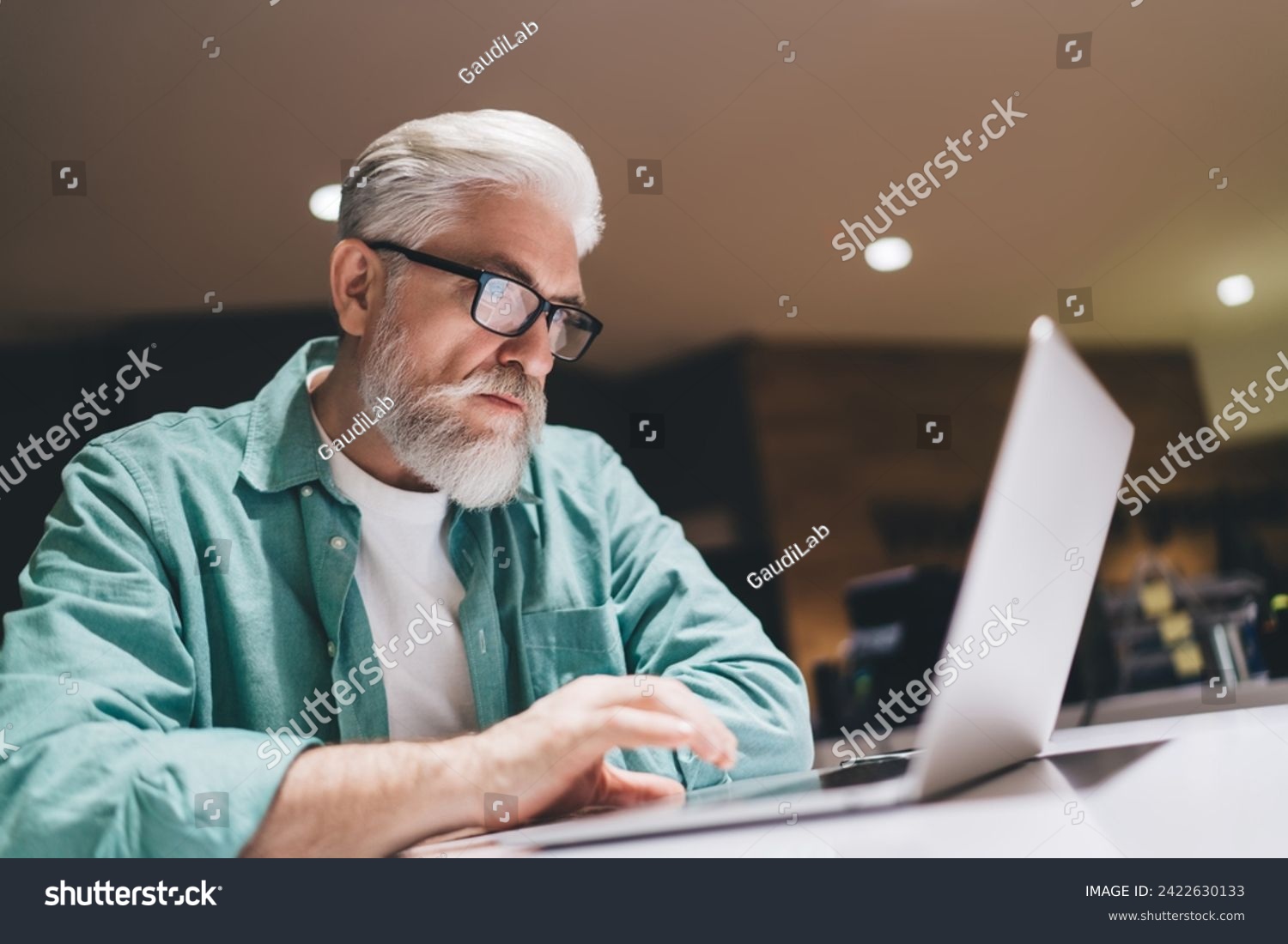 Focused senior Caucasian man with a beard working intently on a laptop in an indoor setting, wearing glasses and a green shirt, symbolizing professional dedication and digital engagement in later life #2422630133