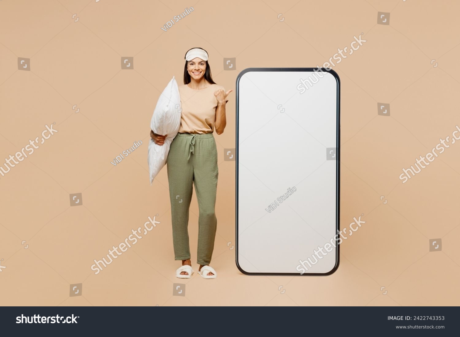 Full body young Latin woman wear pyjamas jam sleep eye mask rest relax point on big huge blank screen area mobile cell phone isolated on plain pastel beige background. Good mood night home nap concept #2422743353