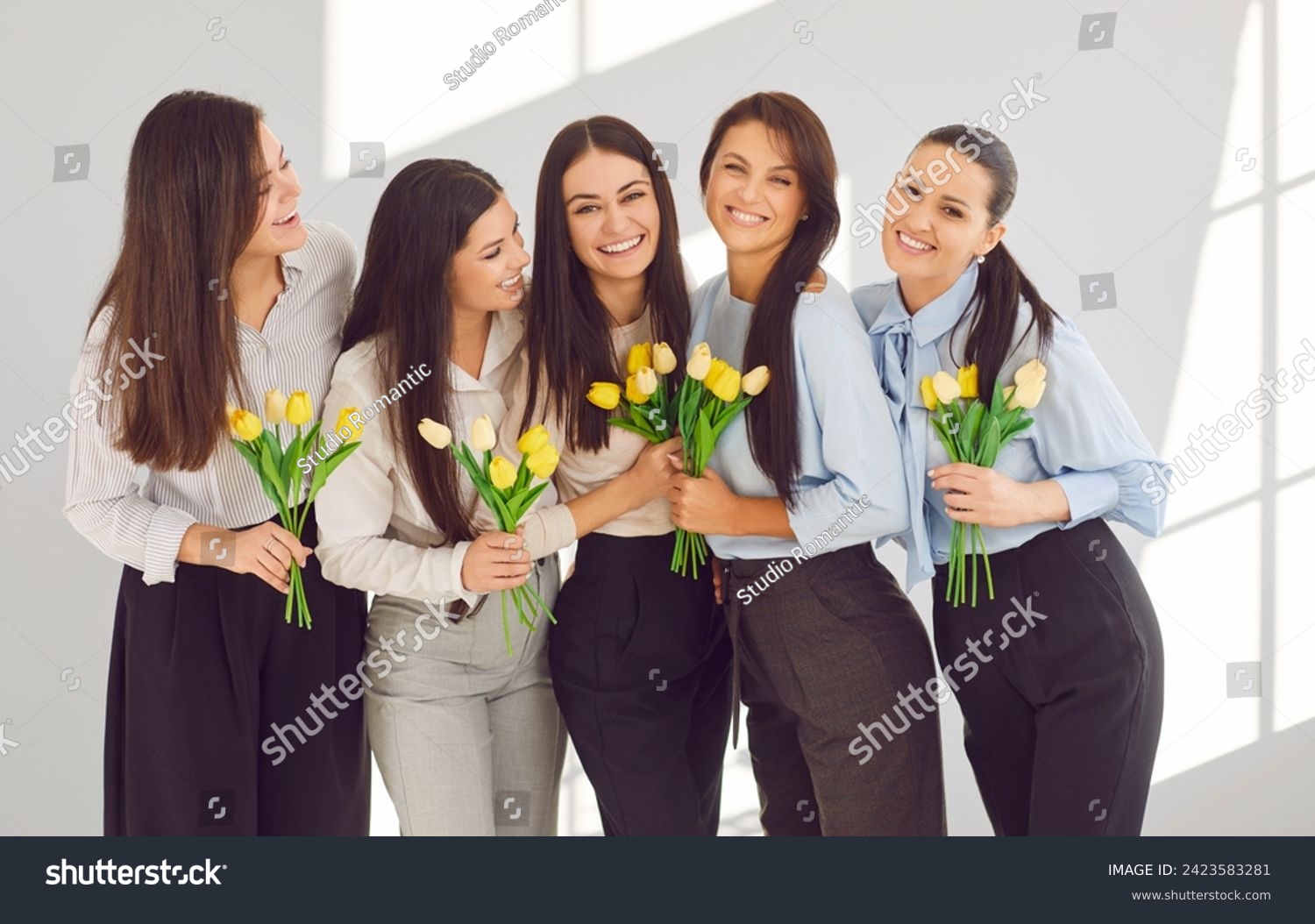 Group of happy business women in office celebrating International Women's Day together. Portrait of stylish and beautiful businesswomen with yellow tulips hugging and smiling in front of camera. #2423583281