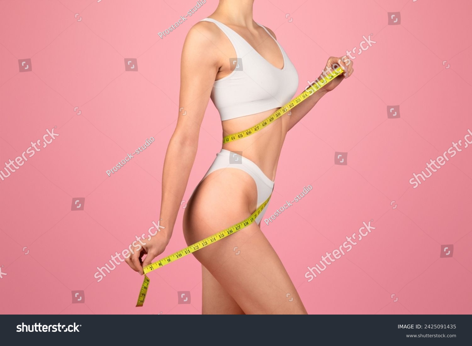 Side profile of a fit woman measuring her waist with a yellow tape measure, dressed in a white sports bra and underwear, against a vibrant pink background, showcasing health and body consciousness #2425091435
