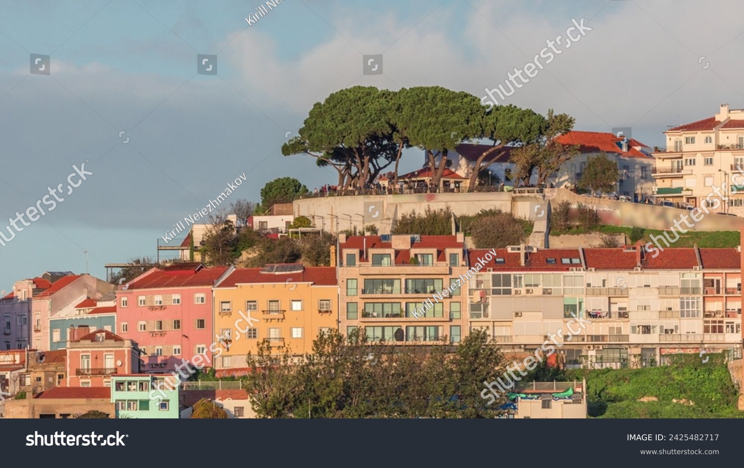 Famous viewpoint Miradouro da Senhora do Monte with city view of Lisbon timelapse during sunset with colorful houses around, Lisbon, Portugal. Clouds on the sky over green trees #2425482717