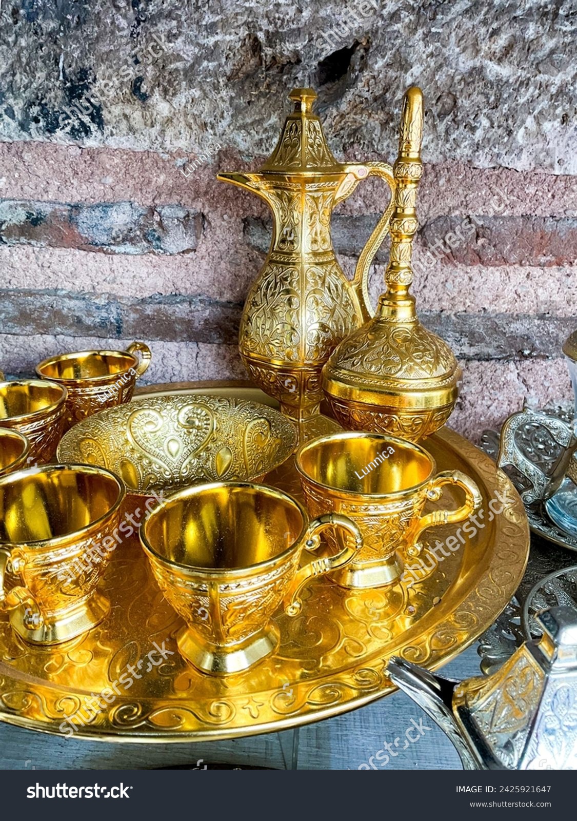 Vintage golden tea set with intricate designs on tray against rustic stone wall. Traditional ornate metalware concept for interior design and decoration. #2425921647