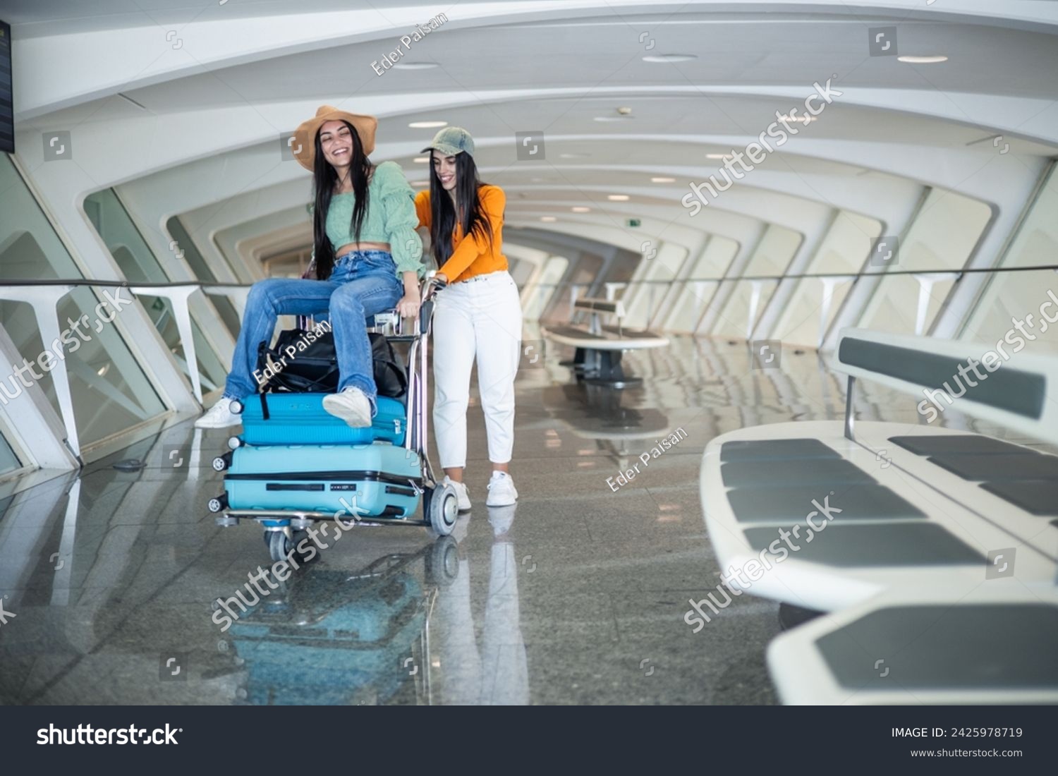 couple of two multicultural girls at the airport sitting on the luggage cart walking having fun #2425978719
