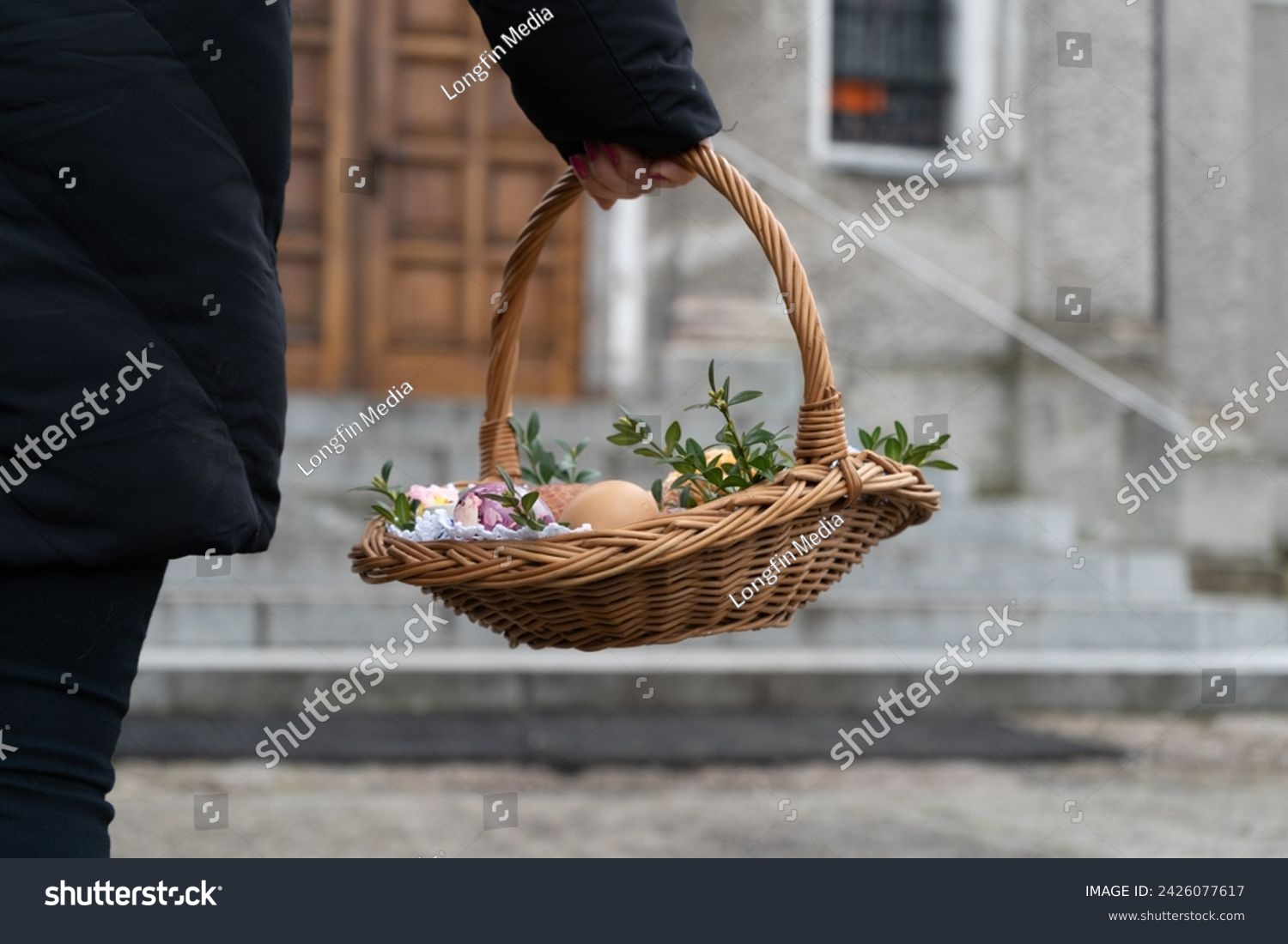 Woman holding Easter basket for blessing in a church. Traditional woven wicker Paschal basket filled with various food, ready to be blessed by a priest as part of the Easter tradition. #2426077617