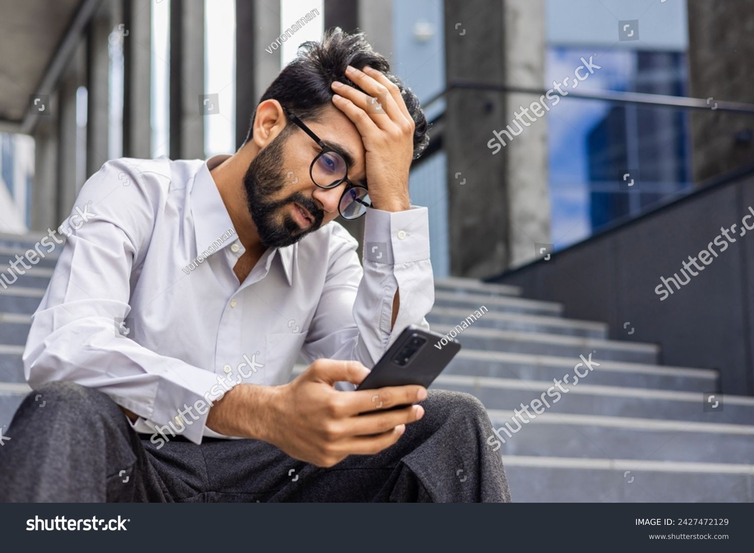 Young man depressed, sad dissatisfied and unhappy outside office building, holding phone, reading bad news from smartphone, businessman in shirt after work. #2427472129