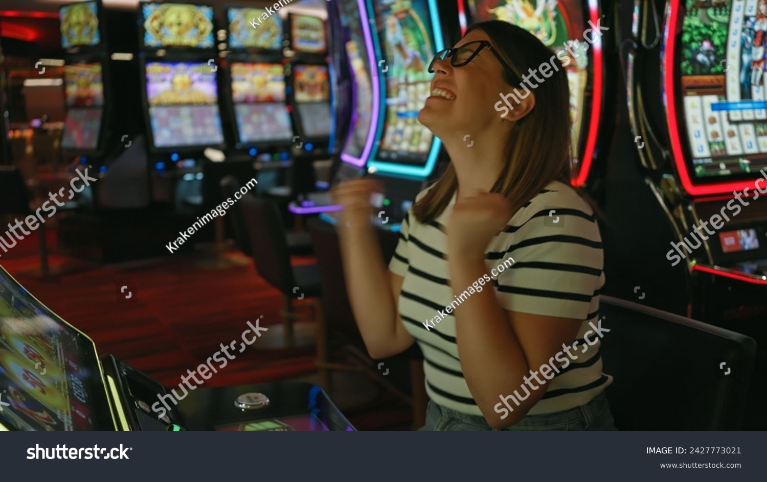 A joyful young adult woman celebrates winning at a casino slot machine. #2427773021