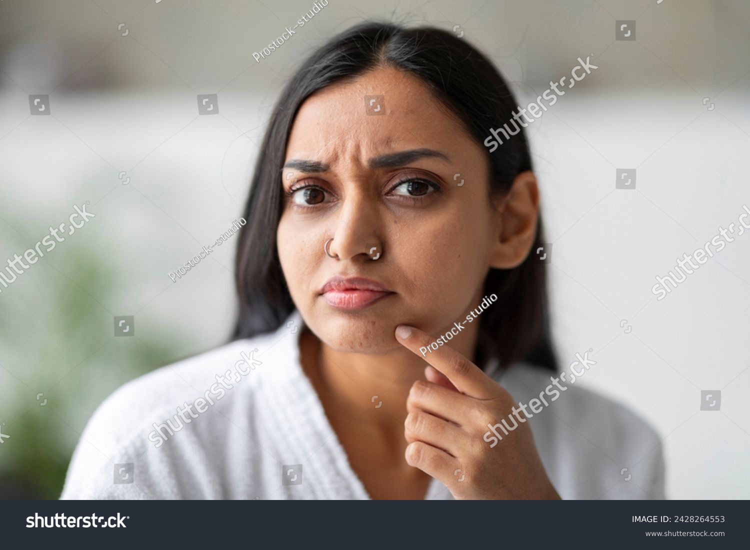 Closeup of upset young indian woman in white bathrobe looking at camera and touching her face at bathroom, suffering from skin problems. Acne, wrinkles, dull skin concept #2428264553