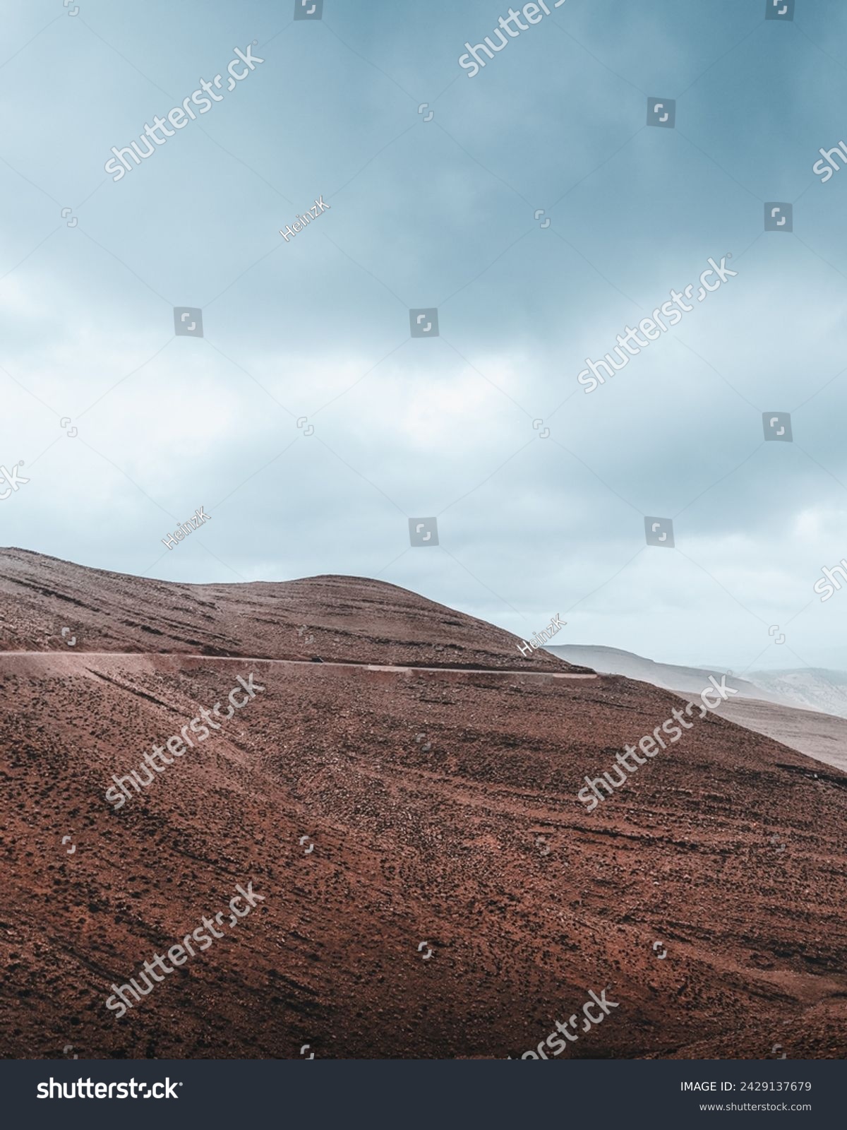 Layers of red and brown earth rise starkly against a dramatic sky, with a winding road cutting through the barren beauty of the Atlas Mountains. The raw, rugged terrain speaks of the earth's deep hist #2429137679