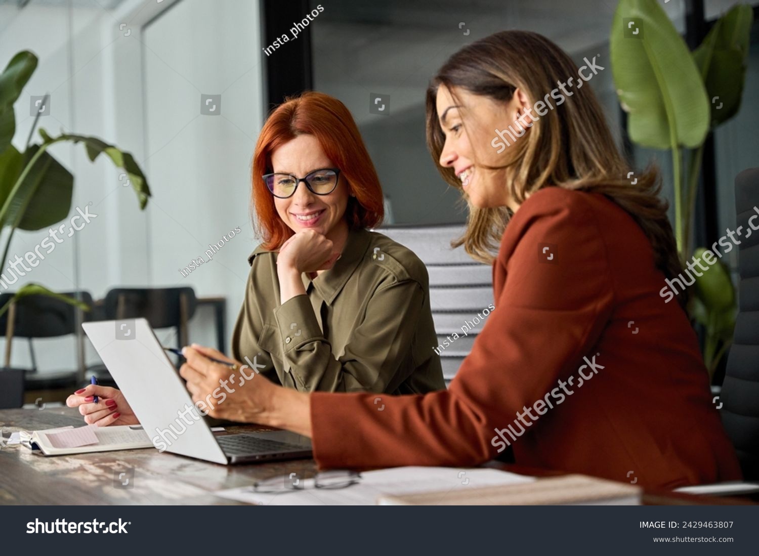 Two happy busy female employees working together using computer planning project. Middle aged professional business woman consulting teaching young employee looking at laptop sitting at desk in office #2429463807