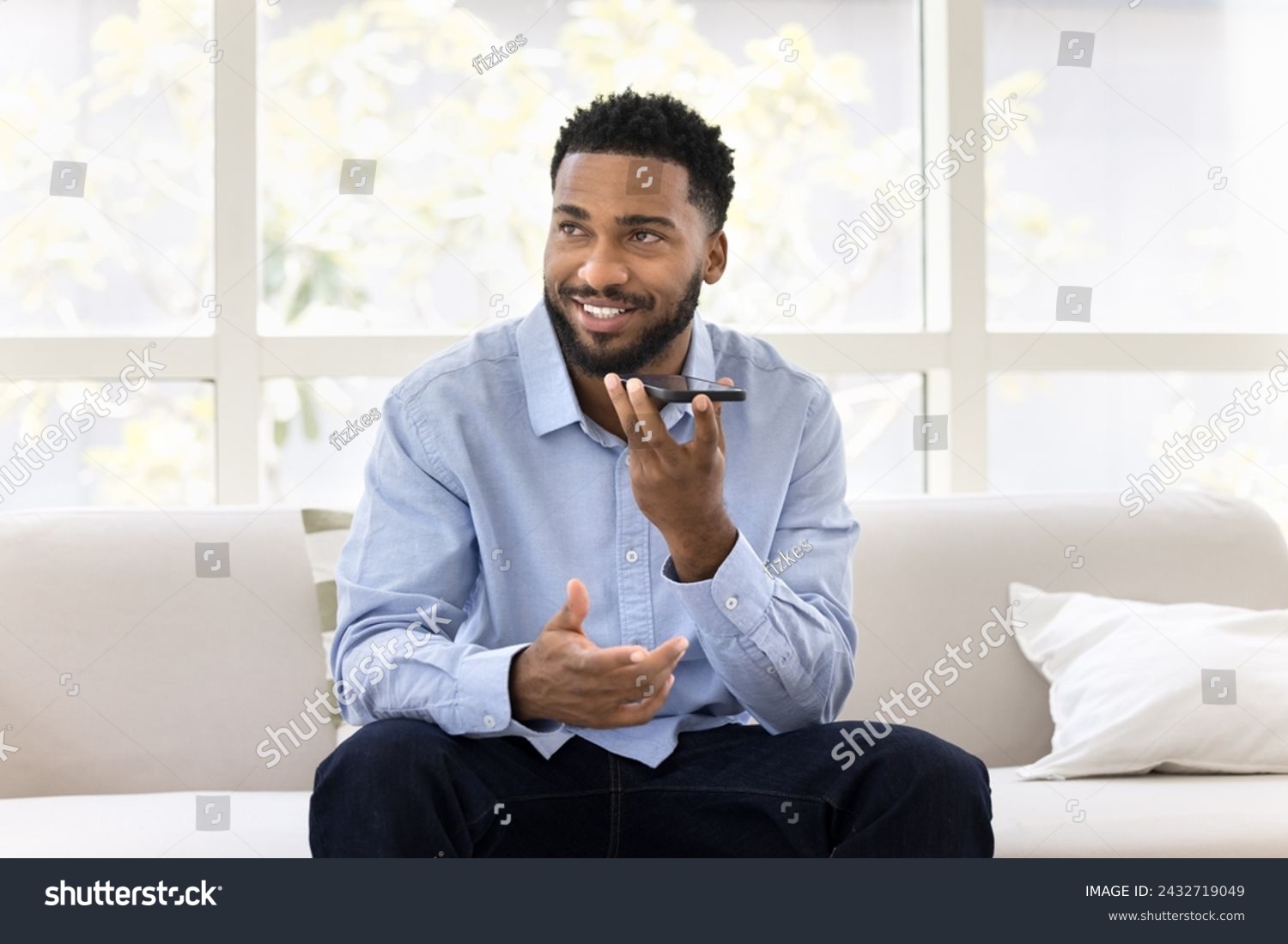 Positive handsome young African business man talking on speaker on cellphone, recording voice message, looking away, thinking, activating modern smart home application #2432719049