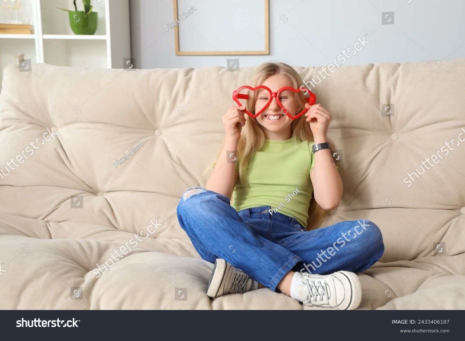 Funny little girl with big eyeglasses sitting on sofa at home. April Fools' Day celebration #2433406187