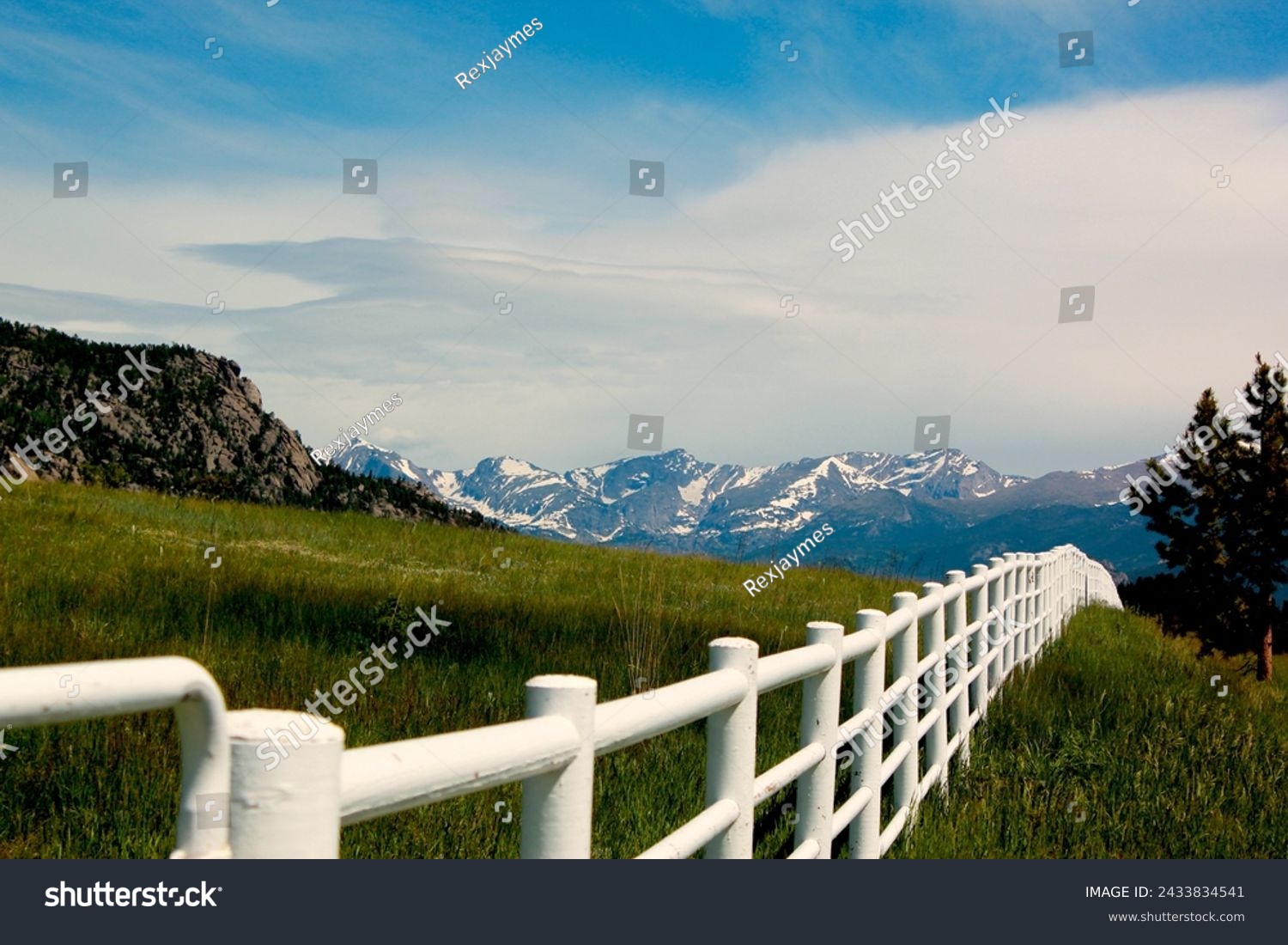 White rail fence with green grass and mountains in background.  Colorado high country scene of rural area.  Blue sky and white clouds. White fence with hills and mountains in the background.   #2433834541