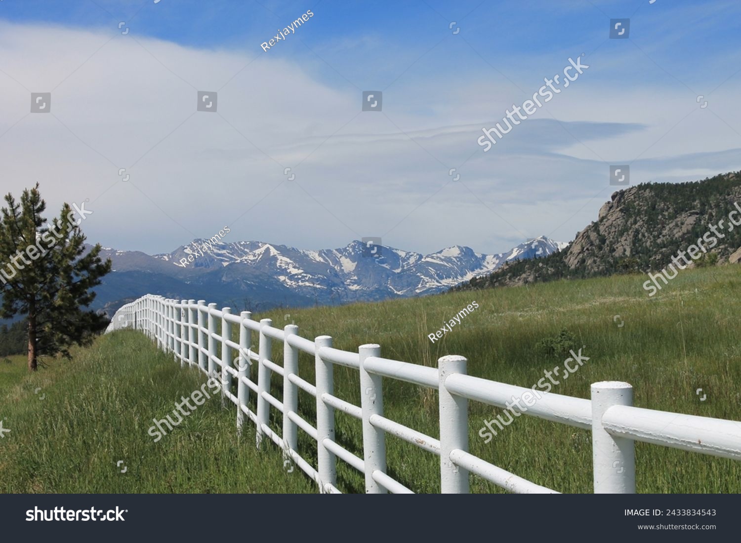 White rail fence with green grass and mountains in background.  Colorado high country scene of rural area.  Blue sky and white clouds. White fence with hills and mountains in the background.   #2433834543