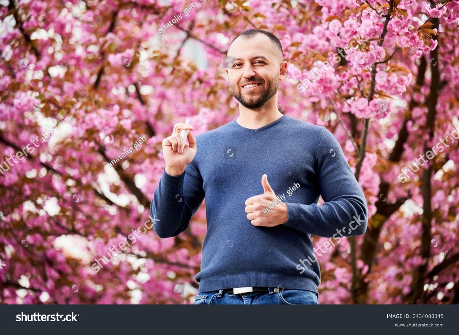 Man allergic using medical nasal drops, suffering from seasonal allergy at spring in blossoming garden. Portrait of smiling man showing thumbs up near blooming tree outdoors. Spring allergy concept. #2434088345
