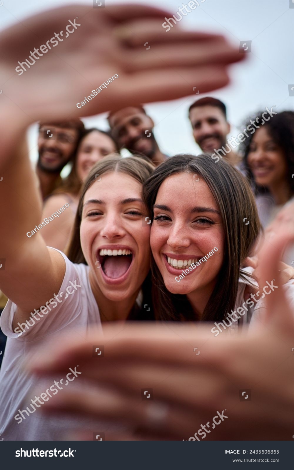 Vertical photo. Two girl friends making a frame with hands. A group of young people is happily celebrating, taking a selfie together. Smiling women having fun during their leisure travel #2435606865