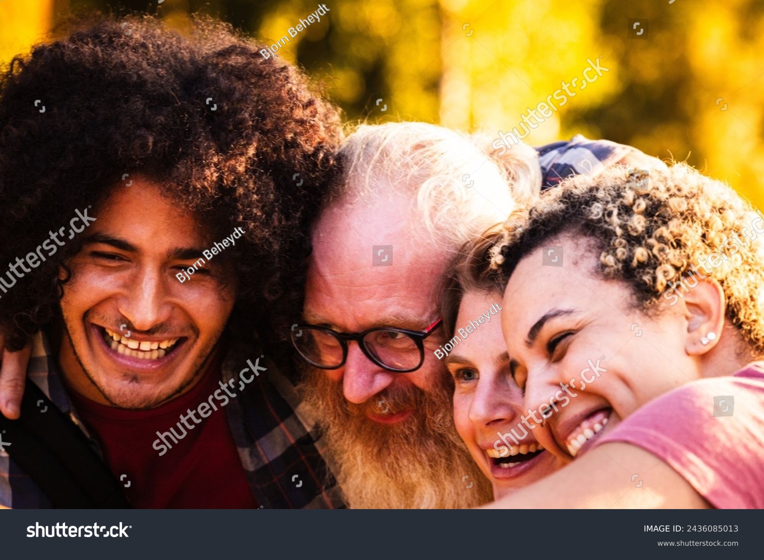 The image captures an up-close, joyful moment among a group of diverse friends. A young man with curly hair is seen laughing alongside an older man with a white beard and glasses, and a young woman #2436085013