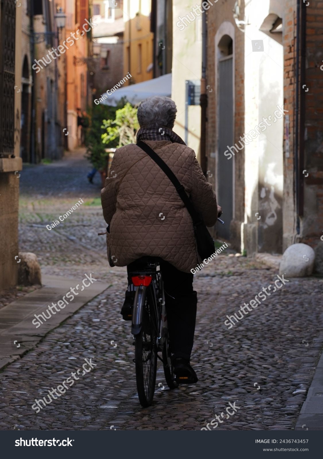 Ferrara, Italy. Elderly woman on a bicycle in a narrow alley in the historic center. #2436743457