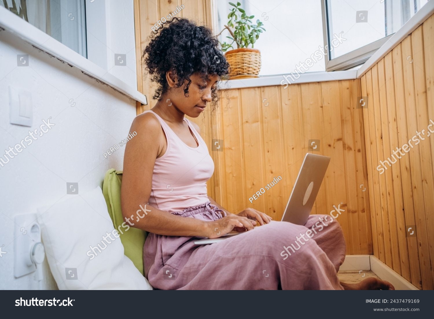 A young woman in casual attire focuses intently on her laptop in a cozy, well-lit wooden alcove. The relaxed home setting emphasizes comfort and productivity. #2437479169