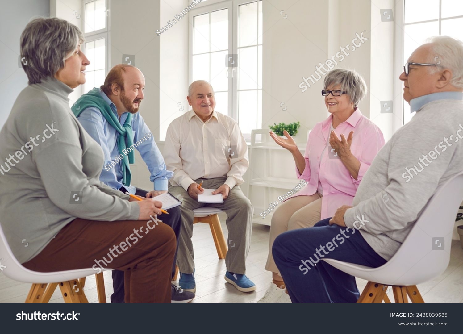 Several old people in group therapy at the retirement home. Senior men and women sitting in a circle, talking about their lives, sharing thoughts and ideas, and taking notes in paper notebooks #2438039685