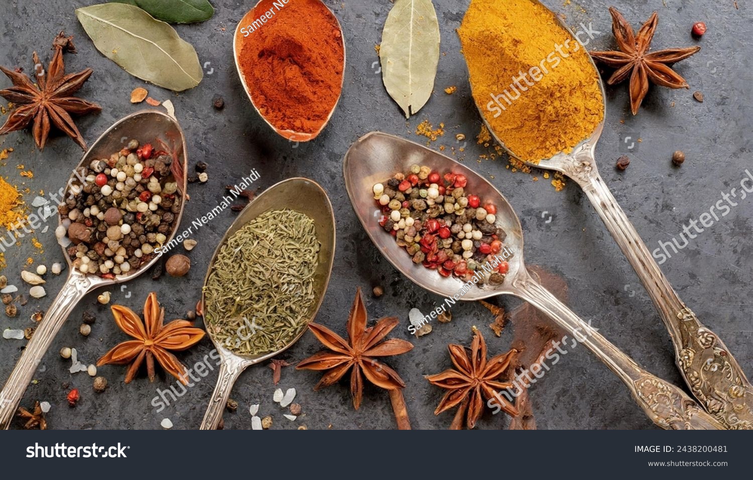 A circle of wooden spoons holding an array of assorted spices rests on a rustic stone tabletop. This vibrant display resembles an exotic Oriental spice market, brimming with colorful herbs and season. #2438200481