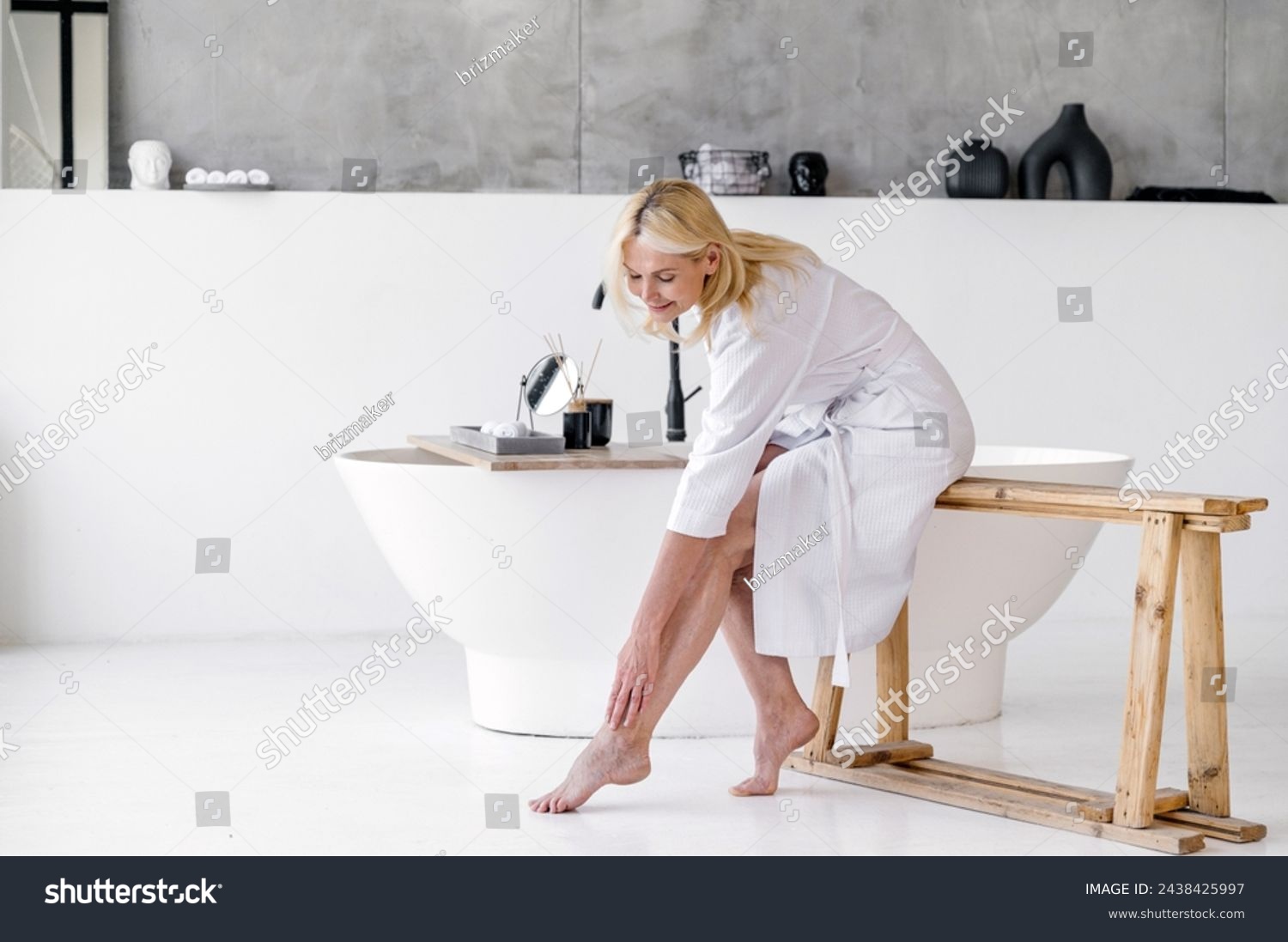 Mid age mature woman applying varicose prevention treatment cream massaging her barefoot legs sitting on bathtub wearing white bathrobe. Concept of health care and bodycare after menopause #2438425997
