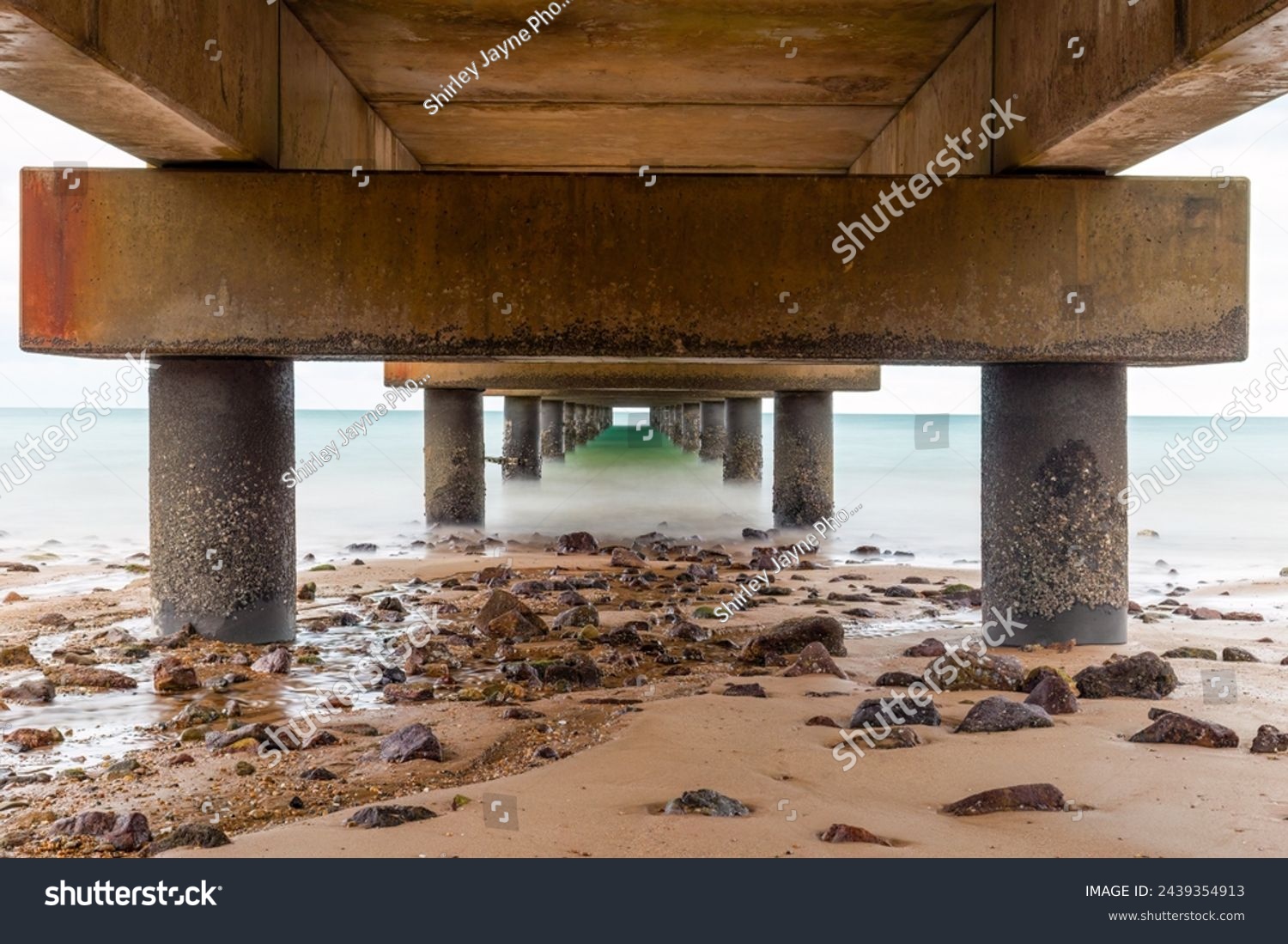 The underneath of a fishing jetty in diminishing perspective view in long exposure showing sand, rocks and support pillars on a tropical beach in Mission Beach in Queensland, Australia. #2439354913