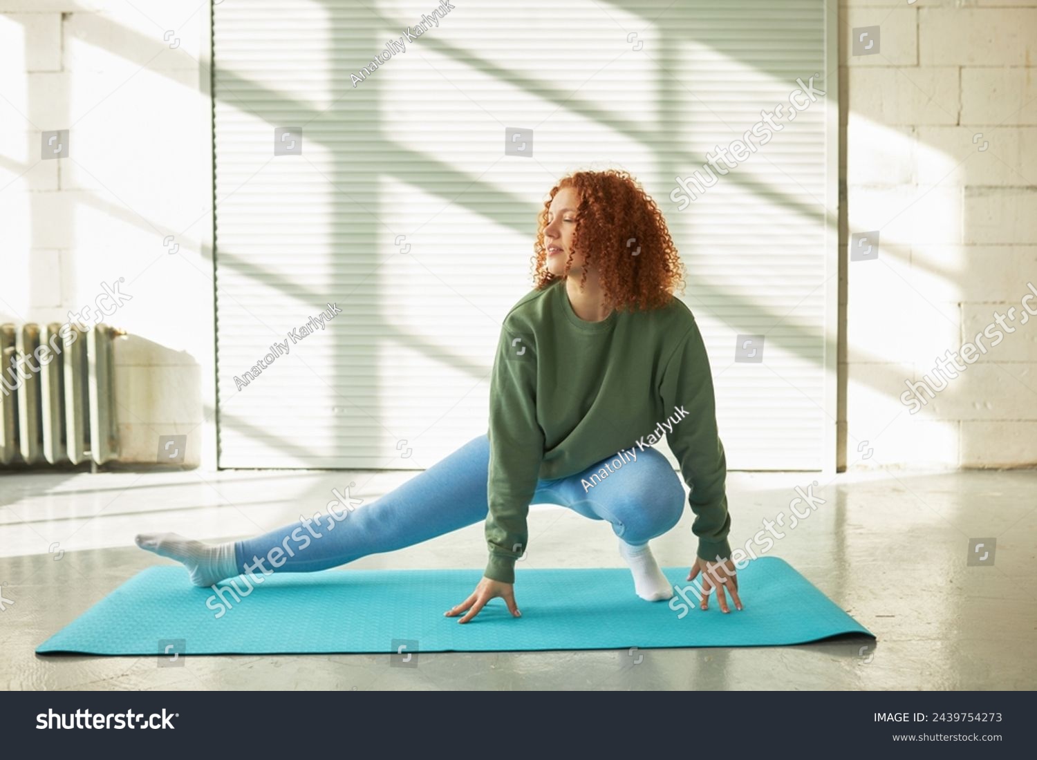 Full-length portrait of flexible redhead woman in sports clothes at gym stretching leg, bending one knee, training indoors, keeping fit and losing weight, enjoying physical exercises #2439754273