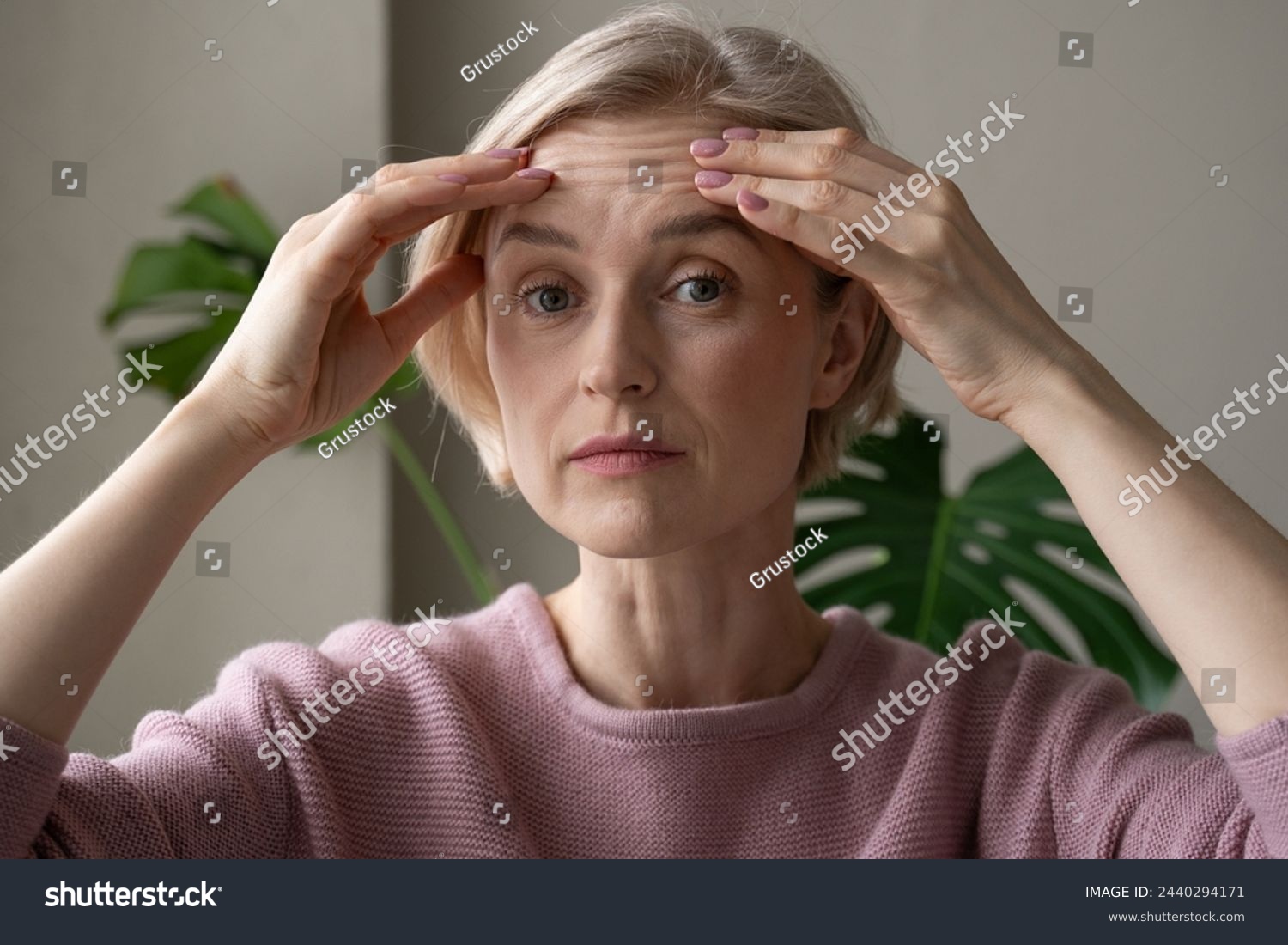 A contemplative mature woman is captured indoors while gently touching her forehead with both hands, appearing introspective or concerned. She wears a soft pink sweater, and her expression conveys #2440294171
