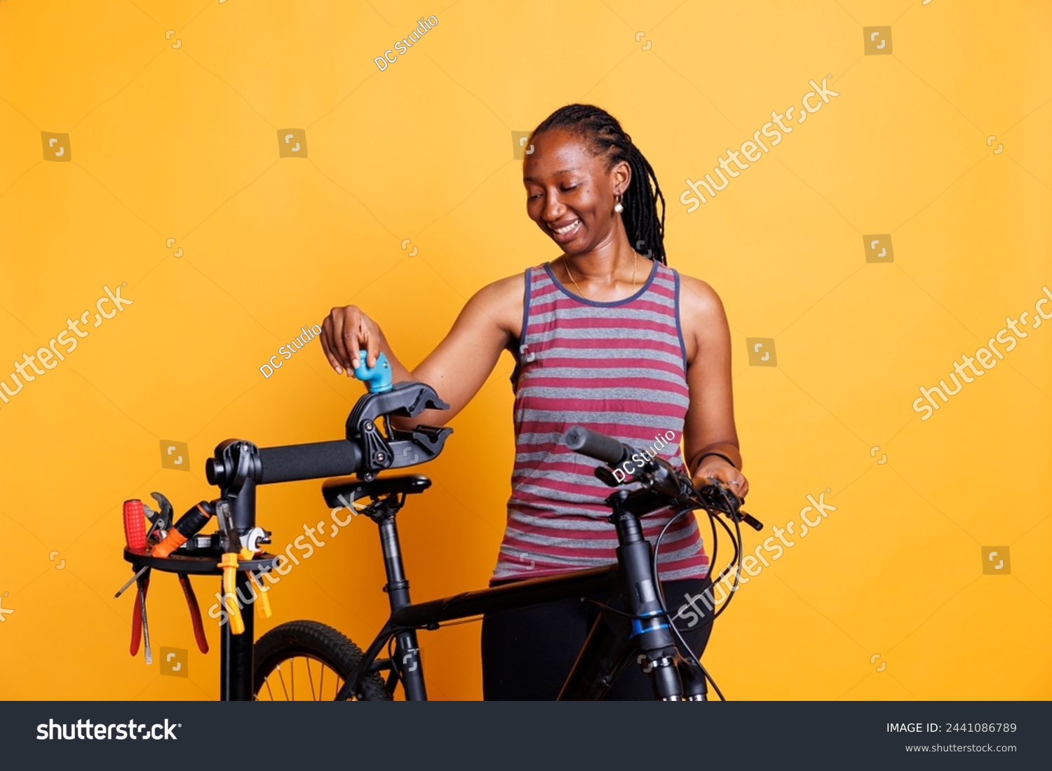 Energetic black woman making bike adjustments with tools and inspecting components. Sports-loving african american lady examining repair-stand for broken bicycle frame. Isolated yellow background. #2441086789
