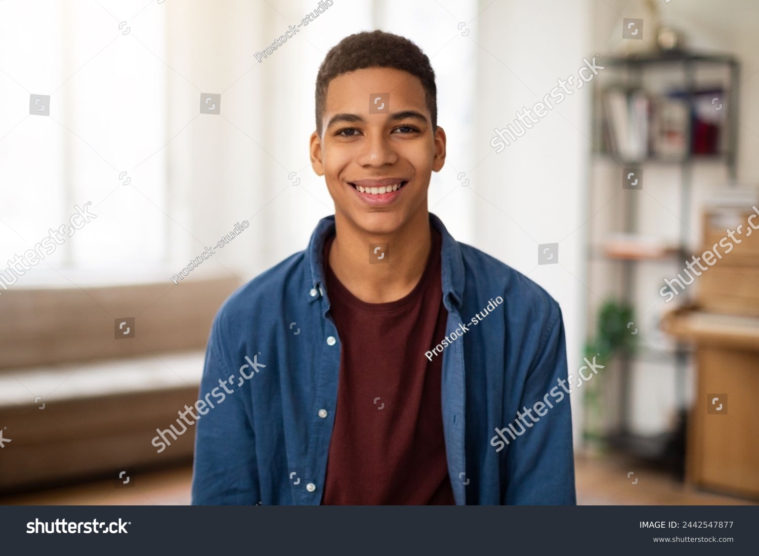 A cheerful young man in a casual shirt and jacket poses with a bright smile in a cozy home environment #2442547877