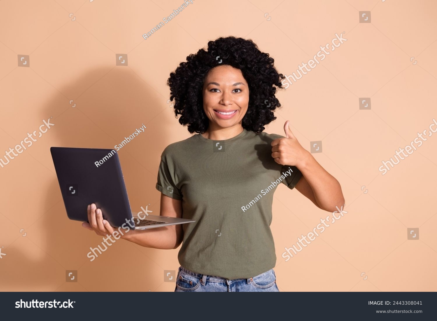 Portrait of multiethnic multinational girl dressed t-shirt hold laptop showing thumb up nice job isolated on beige color background #2443308041