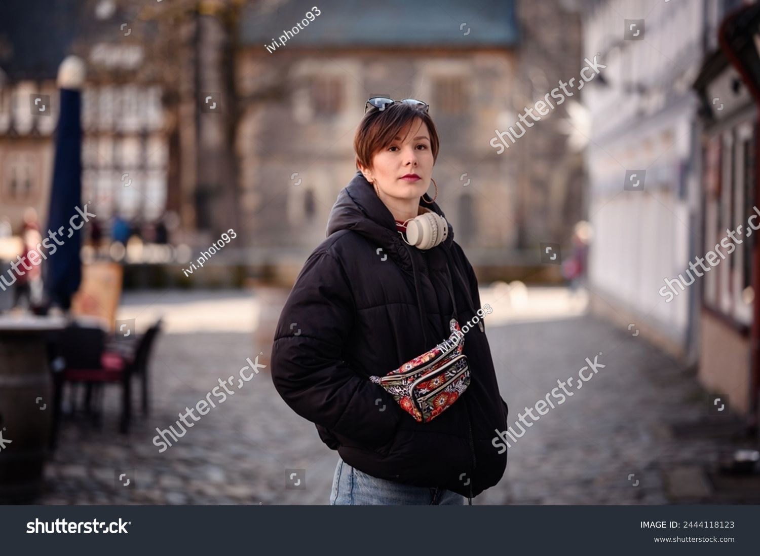A poised adult woman with short hair and red lipstick stands confidently outdoors. She sports casual attire with a patterned fanny pack and headphones around her neck,  #2444118123