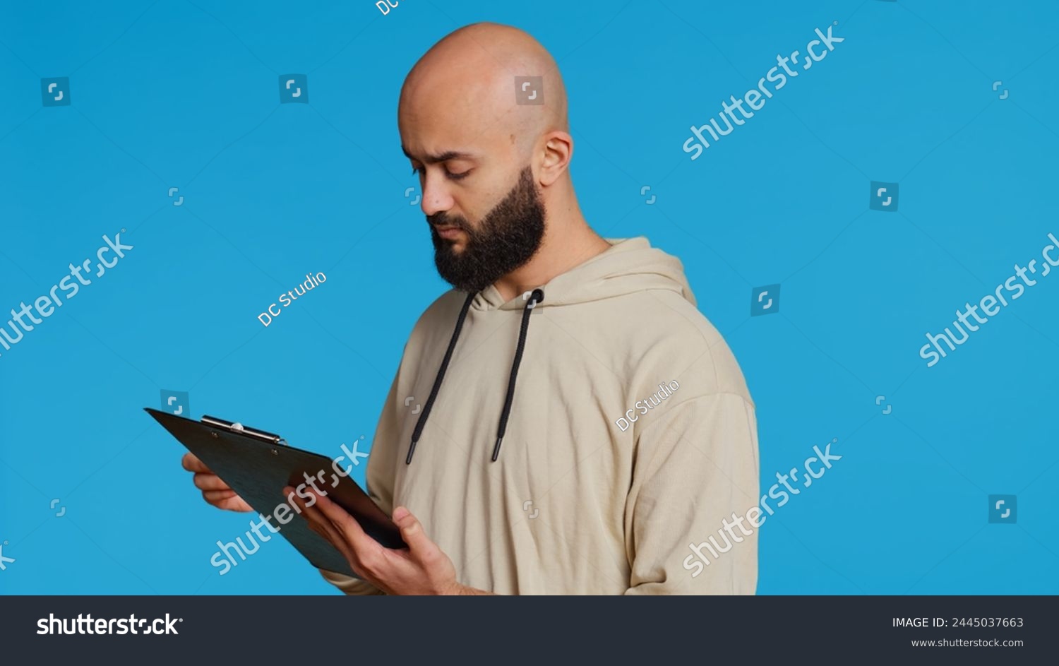 Middle eastern adult creating a checklist on clipboard papers, being focused on trying to plan tasks on an organizer in studio. Modern person looking at files and listing things to do. Camera 2. #2445037663