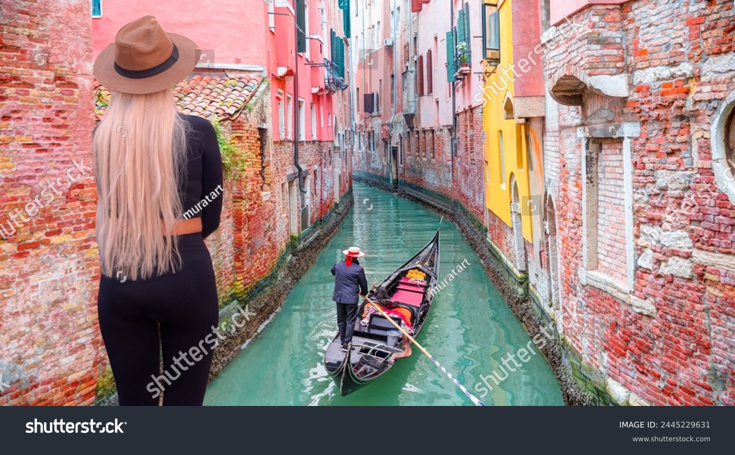 A stylish blonde woman on the bridge watching the Venetian gondolier - Venetian gondolier punting gondola through green canal waters of Venice Italy #2445229631