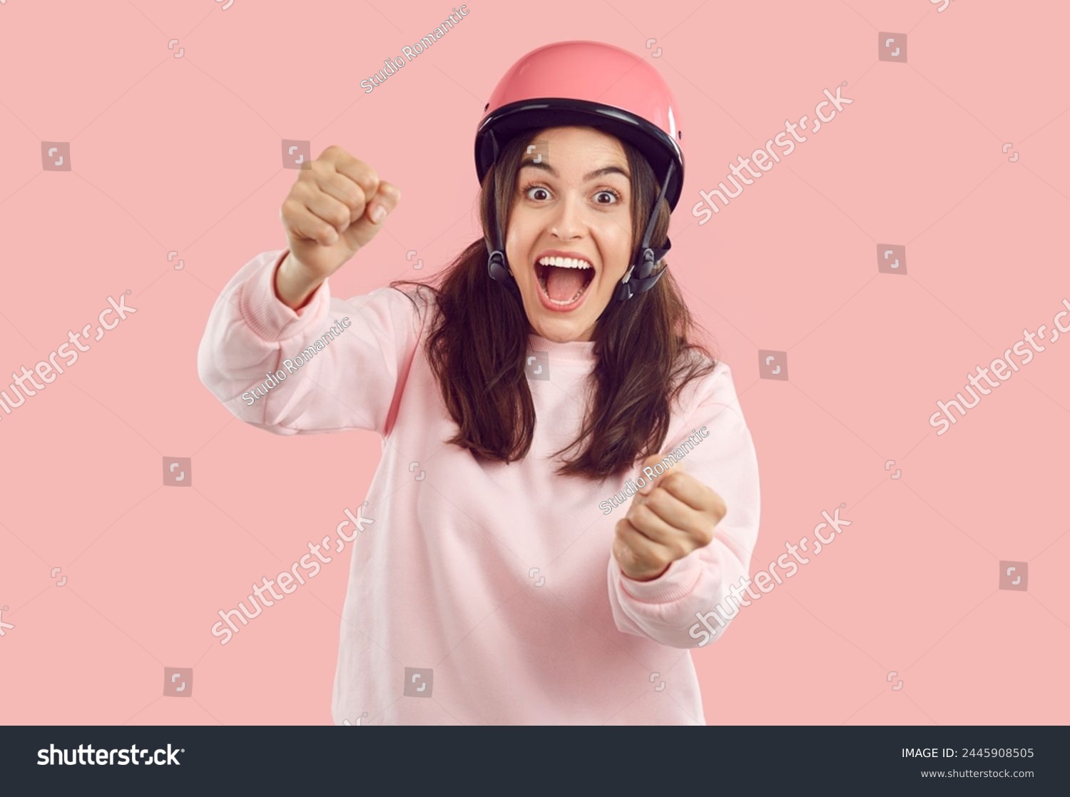 Crazy smiling amazed woman with opened mouth in helmet imagines she is driving and holding steering wheel on pink background. She is wearing pink sweatshirt. Human emotions, energy, drive concept. #2445908505