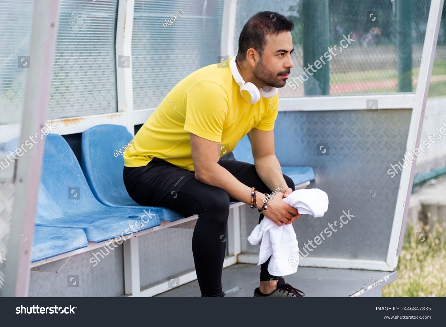 In the arbor of the city sports stadium, a bearded sportsman with headphones and a towel sits on a plastic chair, taking a well-deserved break on the outdoor athletic arena.  #2446847835