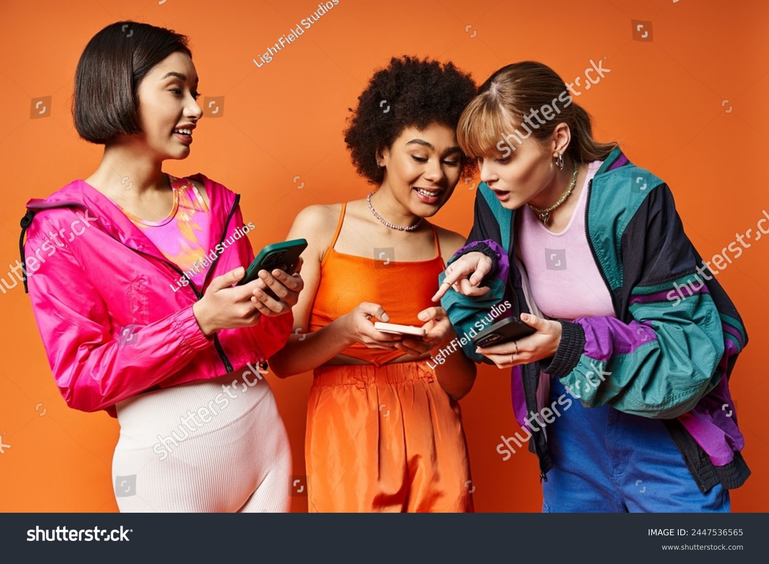 Three diverse women with different ethnicities standing next to each other, absorbed in their cell phones against an orange background. #2447536565