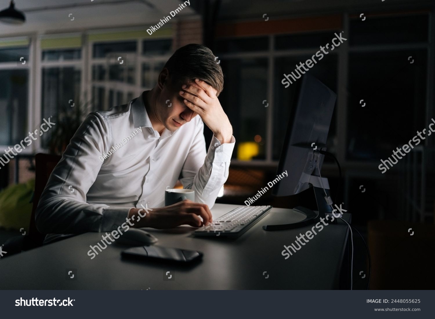 Tired male office worker working late at night, using desktop computer and touching forehead. Exhausted businessman working on laptop computer until night. Concept of stressful life and deadline. #2448055625