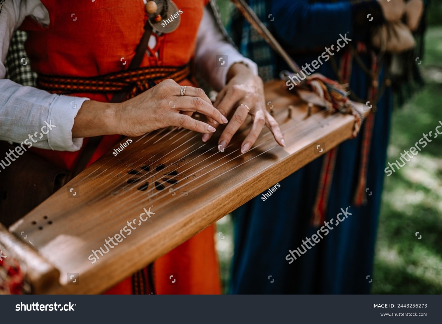Valmiera, Latvia - July 14, 2023 - Hands playing a kokle, a traditional Latvian stringed musical instrument, with musicians in folk costumes blurred in the background. #2448256273