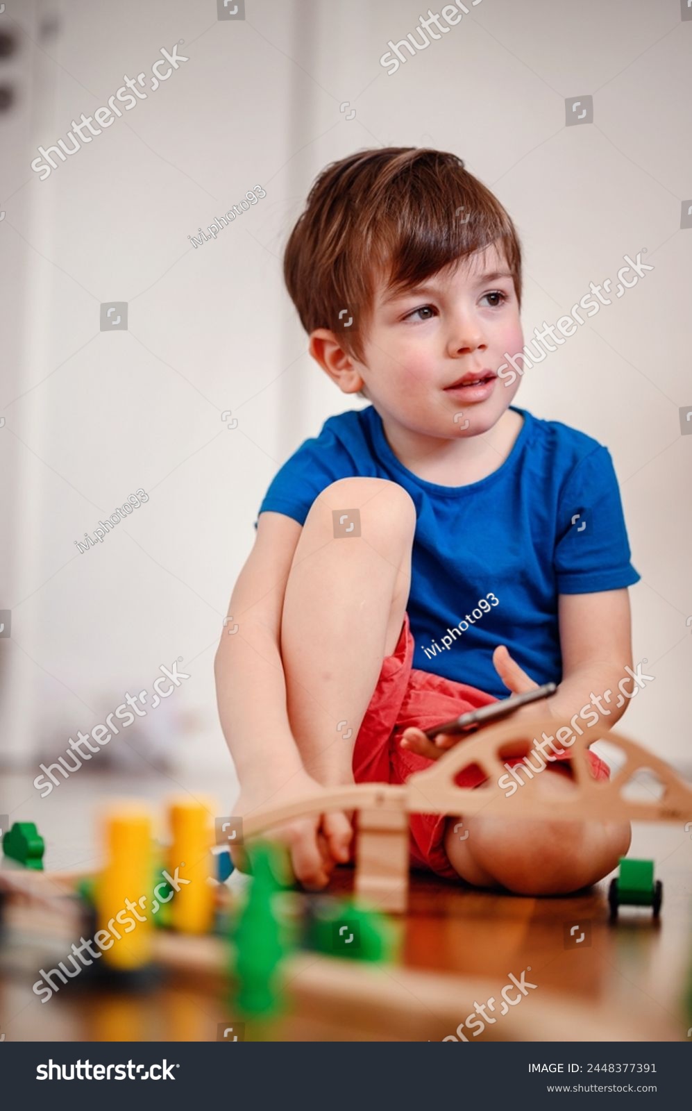 A joyful young boy with a smartphone sits amidst a wooden train setup, his bright laughter echoing the playful fusion of digital and tangible play #2448377391