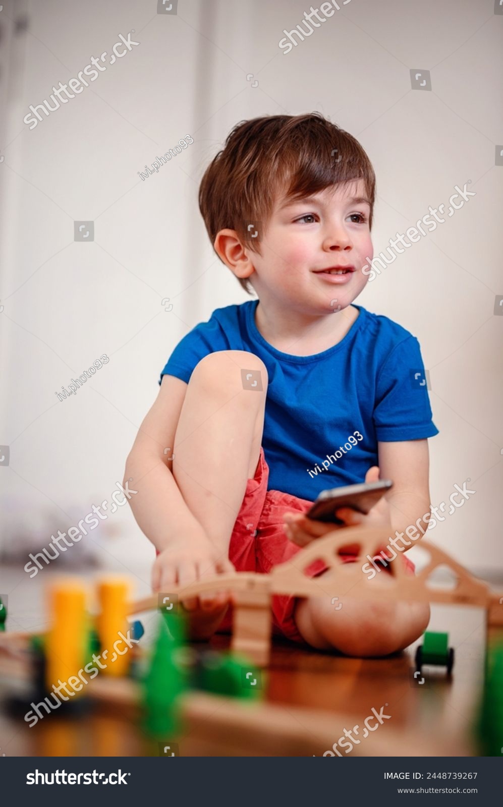 A joyful young boy with a smartphone sits amidst a wooden train setup, his bright laughter echoing the playful fusion of digital and tangible play #2448739267