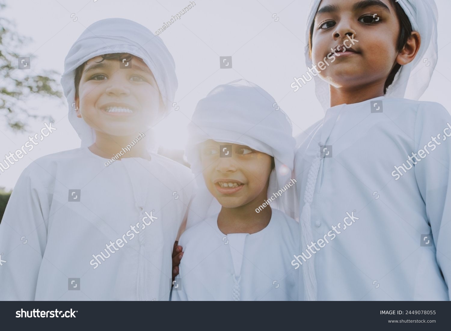 Children playing together in Dubai in the park. Three children with dish dasha look into the camera smiling. Emirati people wearing traditional kandura white dress. #2449078055