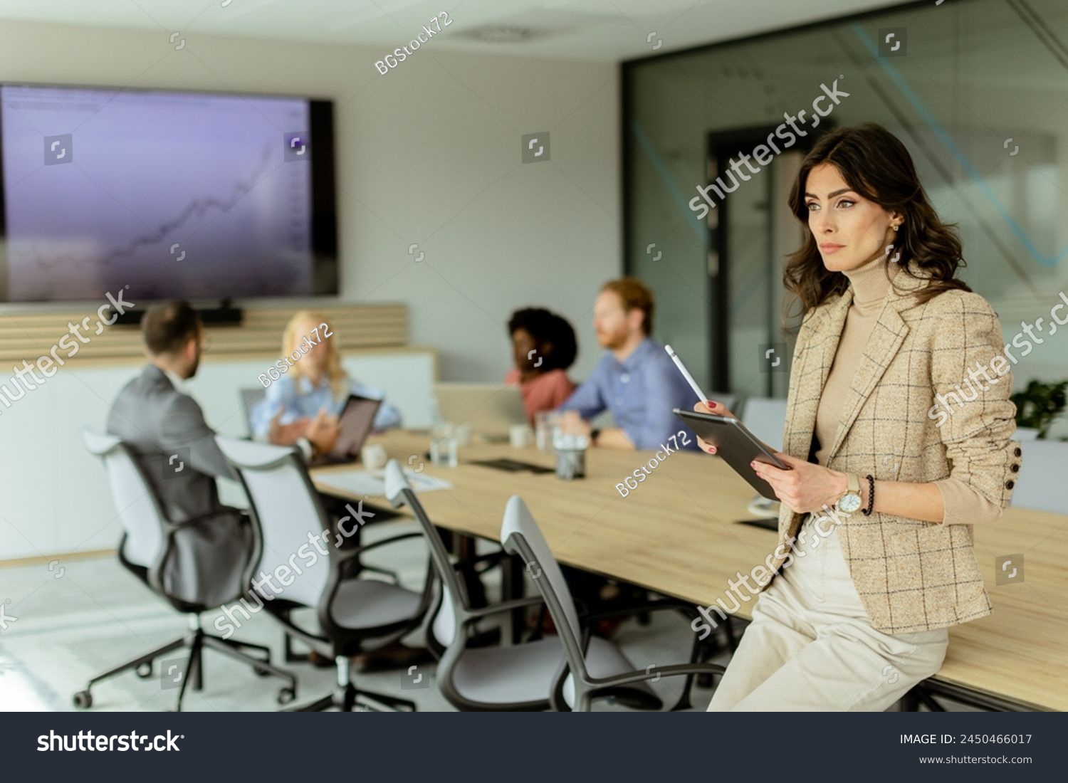 A poised woman with a tablet stands at the forefront of a collaborative office space, her team engaged in discussion behind her. #2450466017