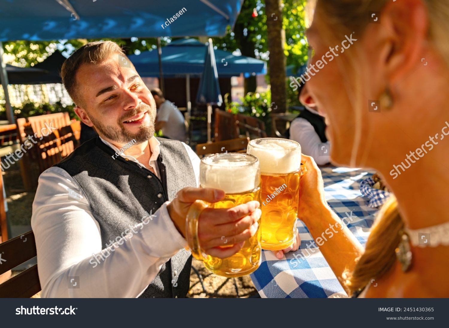 smiling man in traditional attire toasting with a woman in tracht at oktoberfest or beer garden in germany #2451430365