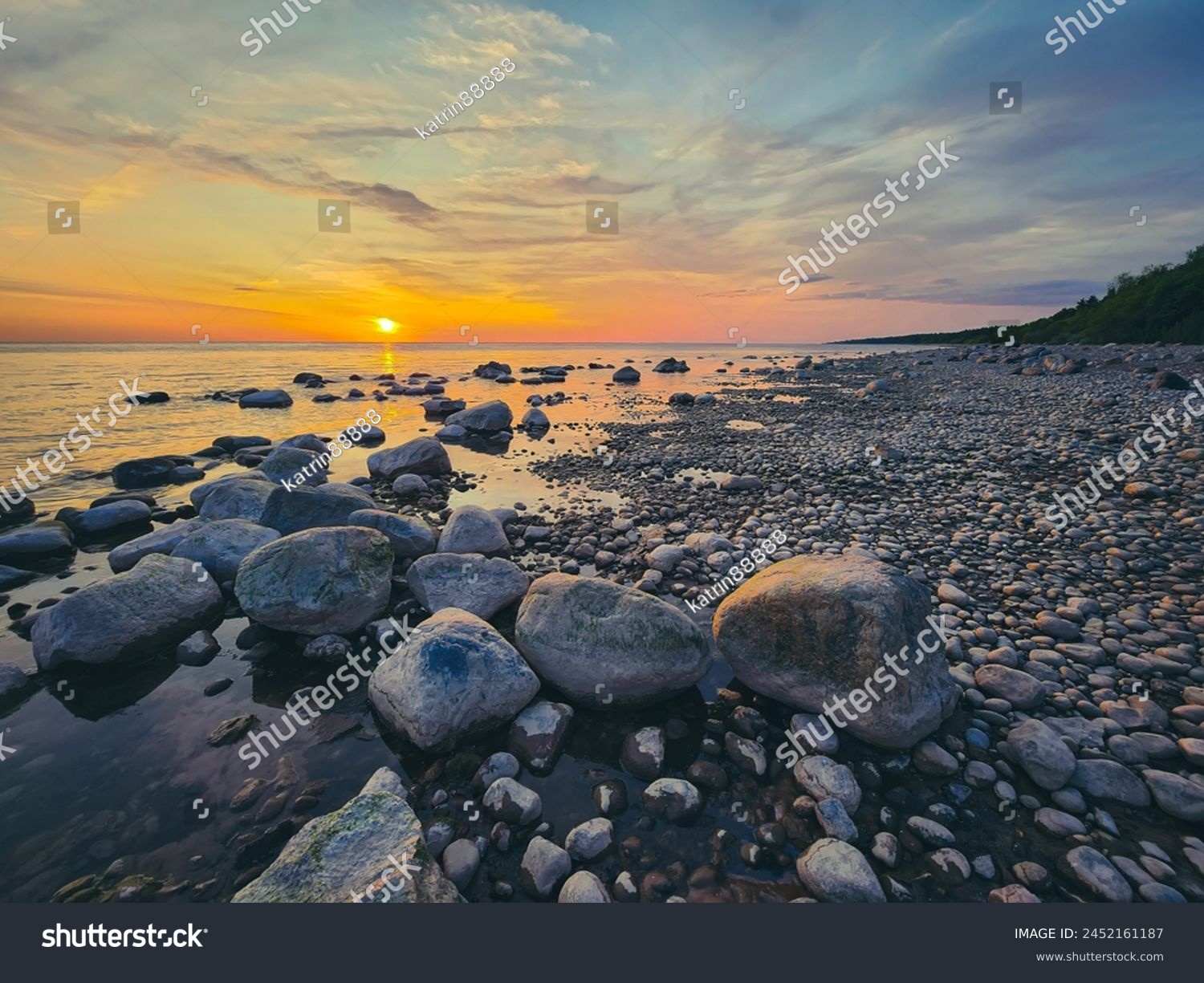 Sunset over serene lakeshore, with calm water reflecting the blue sky. Moss-covered rocks of varying sizes and shapes add texture, while the warm glow of the setting sun creates a tranquil atmosphere. #2452161187